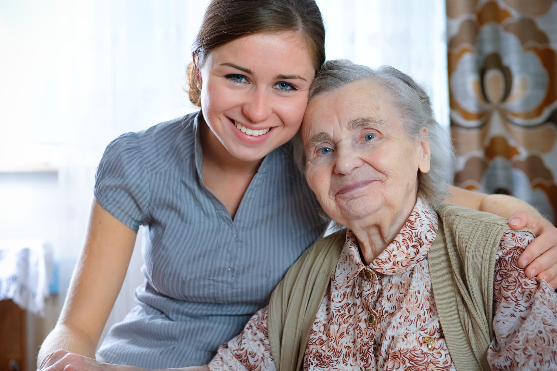 Caregiver sitting cheek to cheek with a smiling senior woman at home